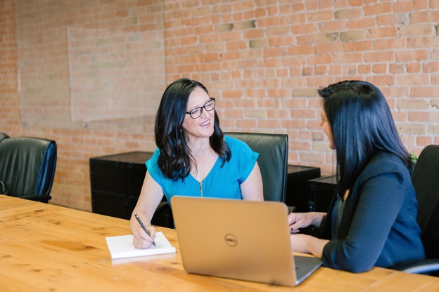 Two women with black hair collaborating at a desk with a notepad, pen and laptop