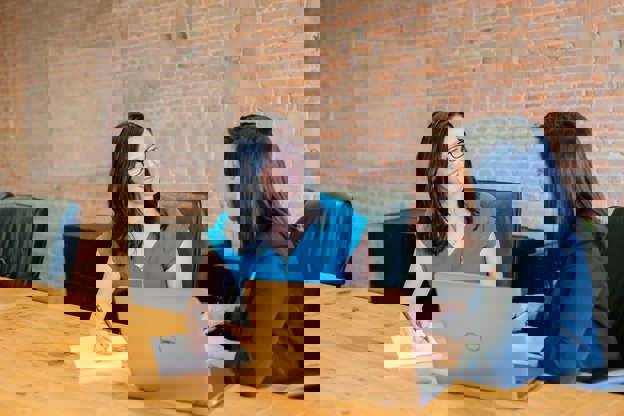 Two women with black hair collaborating at a desk with a notepad, pen and laptop